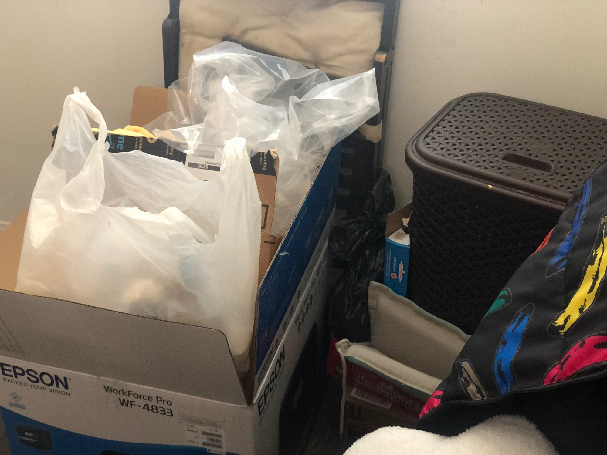 photo of an empty printer box, Epson WorkForce Pro WF-4833, with empty bags and boxes stacked on top, beside a black folding chair with a white cushion, next to a brown plastic trash can with a latticed top. a black jacket with differently colored bananas on a chair in the foreground above something white and fluffy. taken at the robledo art, strike! headquarters, february 2023.