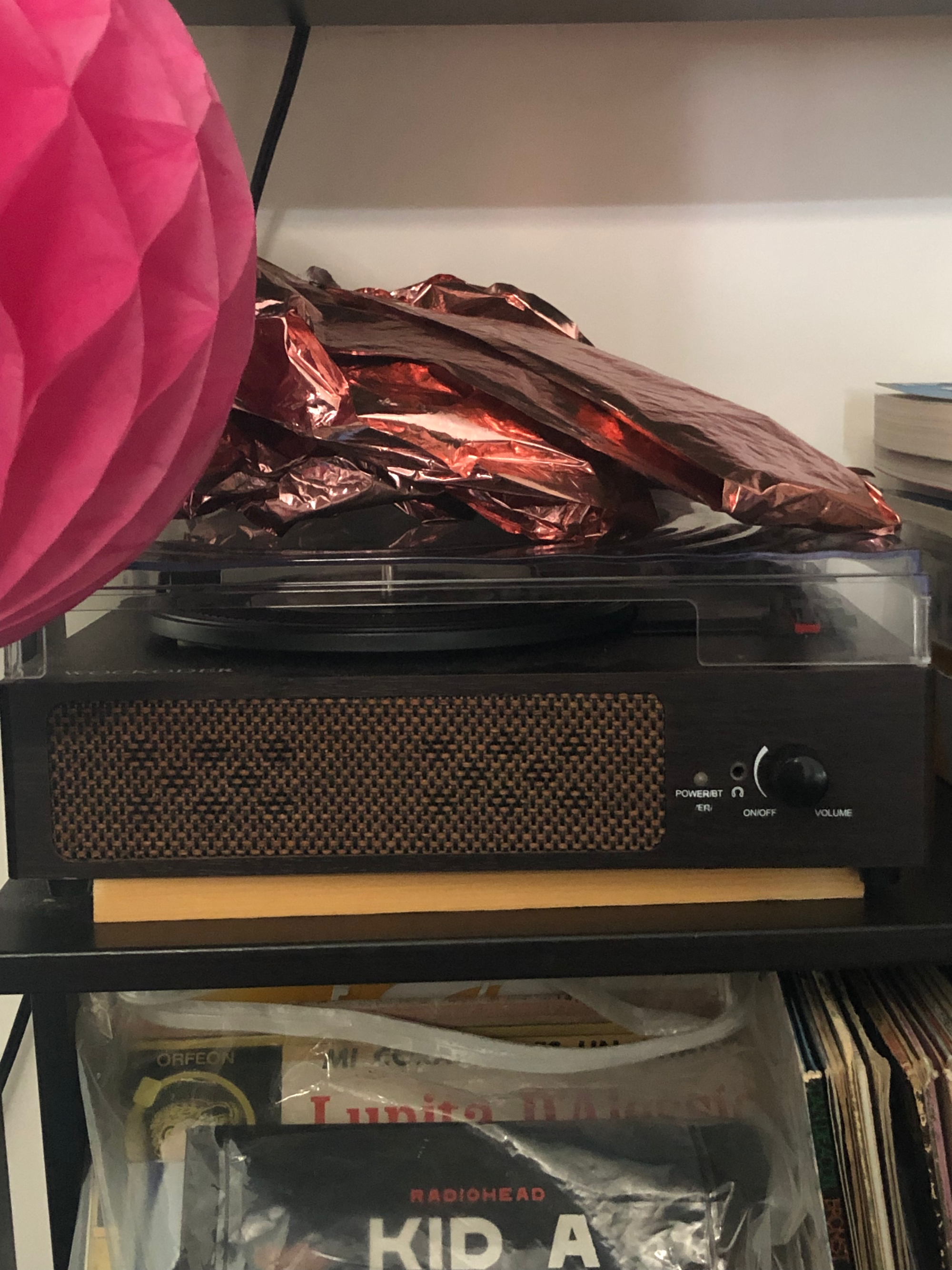 photo of a bookshelf with a dark brown bluetooth-enabled record player sitting on top of a book, the spine facing away from the camera, copper-colored deflated balloon letters on top. on the bookshelf hangs a pink paper orb; on a lower shelf, a bag of records, the first two Kid A by Radiohead and an album by Lupita D'Alessio. taken at the robledo art, strike! headquarters, february 2023.