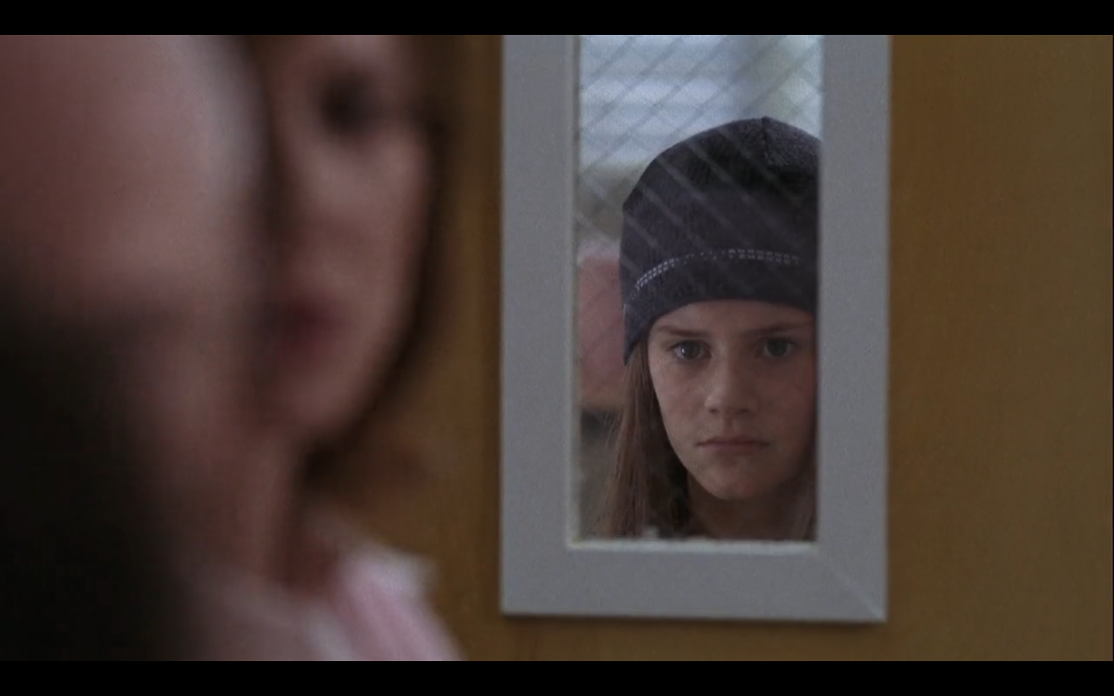 Still from episode 22 of Grey's Anatomy. Bex, a white teenager wearing a beanie, looks out at their parents through the gridded rectangle of an exam room window.